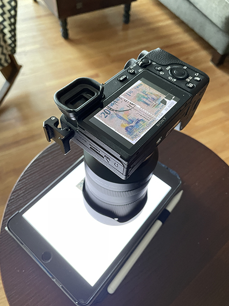 A Sony mirrorless camera with a Tamron lens positioned over a backlit postage stamp on an iPad&mdash;demonstrating how to photograph stamps using natural lighting and macro setup for high-detail results.