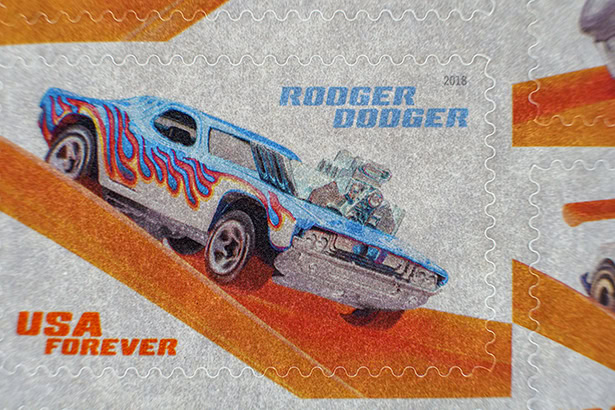 A vibrant close-up of a 2018 USA Forever stamp featuring the Hot Wheels car "Rodger Dodger" with blue body paint and red-orange flames on a stylized ramp background. &copy;Jillian Bell