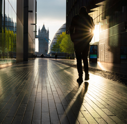 Silhouette of a person walking through a modern urban walkway at sunset, with Tower Bridge visible in the background.