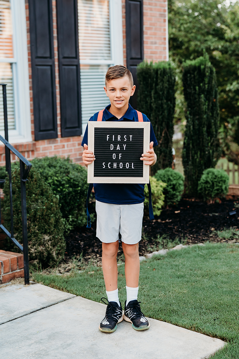 Jeune garçon debout sur un trottoir devant une maison, tenant une pancarte sur laquelle on peut lire "Premier jour d'école". Il est vêtu d'un polo marine, d'un short clair et de baskets, avec un sac à dos bleu, prêt pour les portraits de la rentrée scolaire.