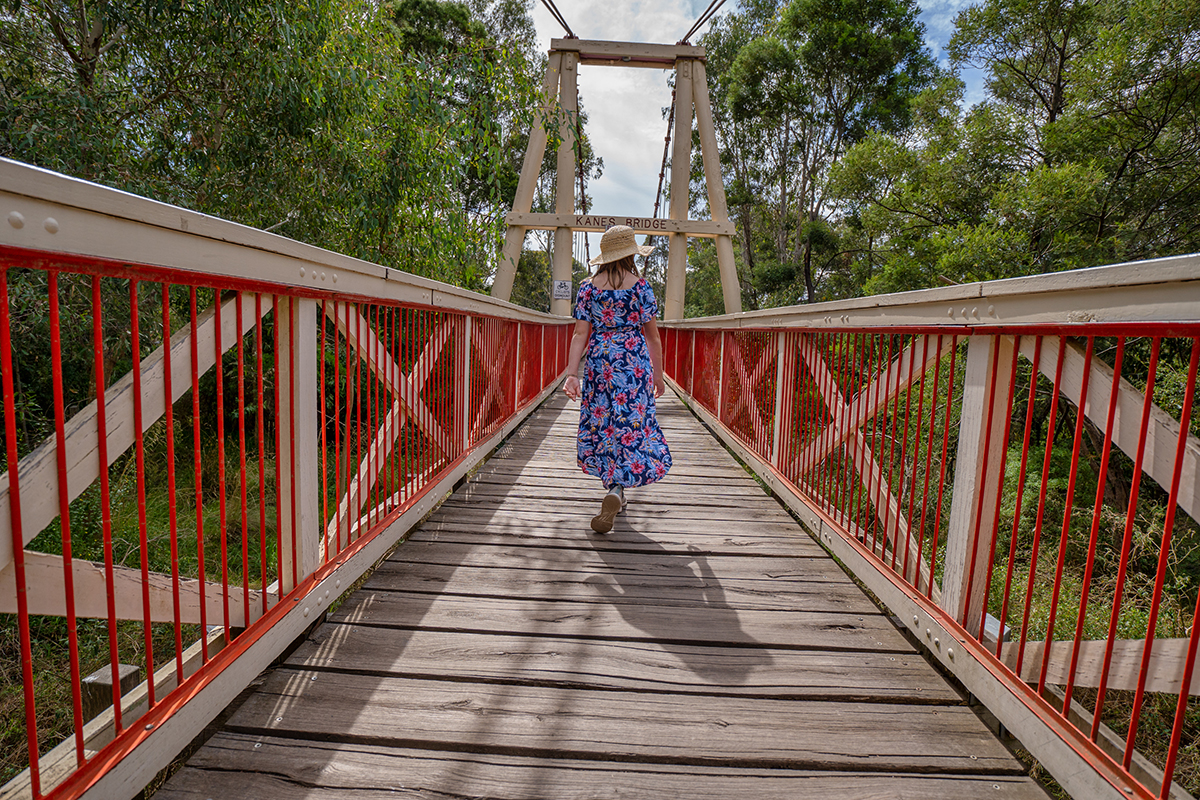Una mujer con un vestido de flores camina por un puente colgante de madera, con fuertes líneas principales creadas por las barandillas rojas y los tablones de madera que guían la mirada del espectador hacia el centro del encuadre.