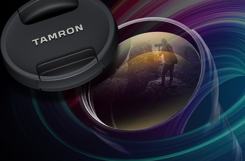Close-up de uma lente Tamron com tampa e reflexo de um fotógrafo no vidro, em um fundo abstrato colorido.