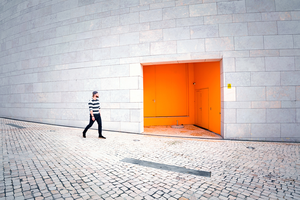 Man in a white and grey striped shirt walking past a vibrant orange door on a gray stone wall.