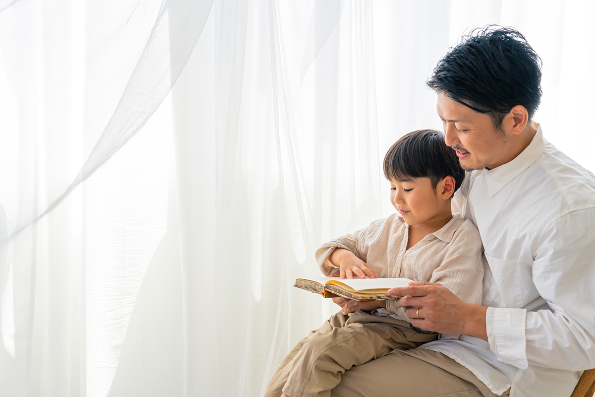 Padre e hijo en atuendo ligero leyendo un libro juntos junto a una ventana luminosa con cortinas blancas.
