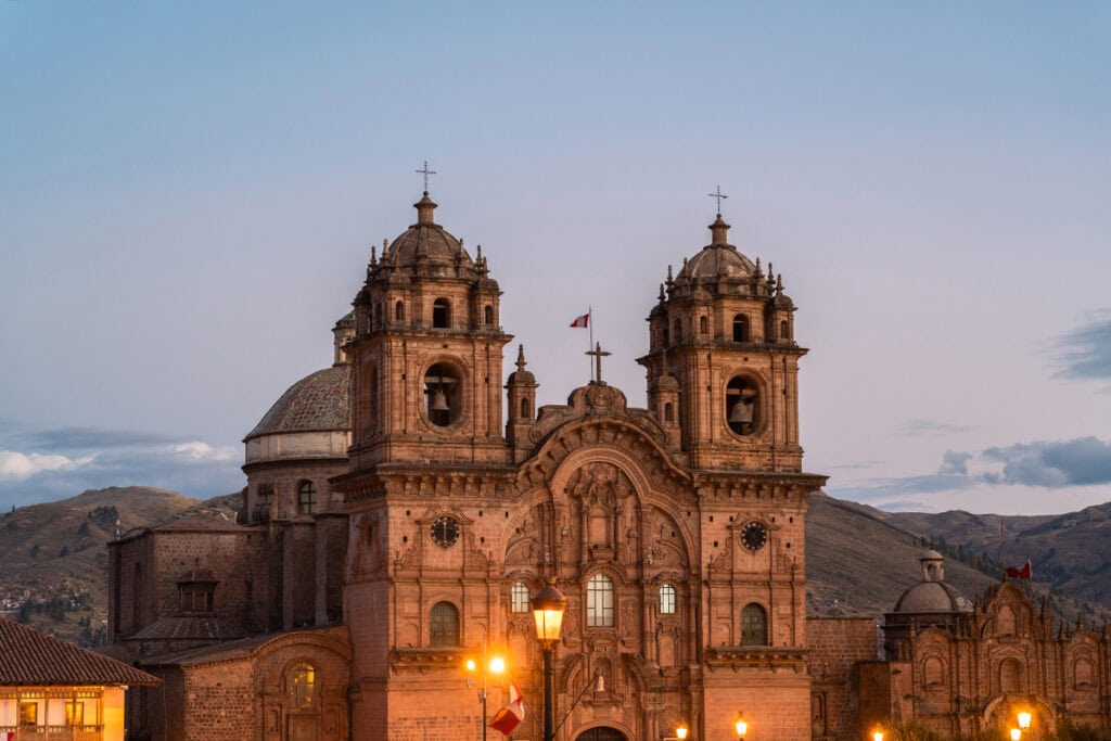 Cathédrale historique au crépuscule, sur fond de montagnes, avec des lumières chaudes illuminant sa façade de pierre détaillée et ses clochers jumeaux.