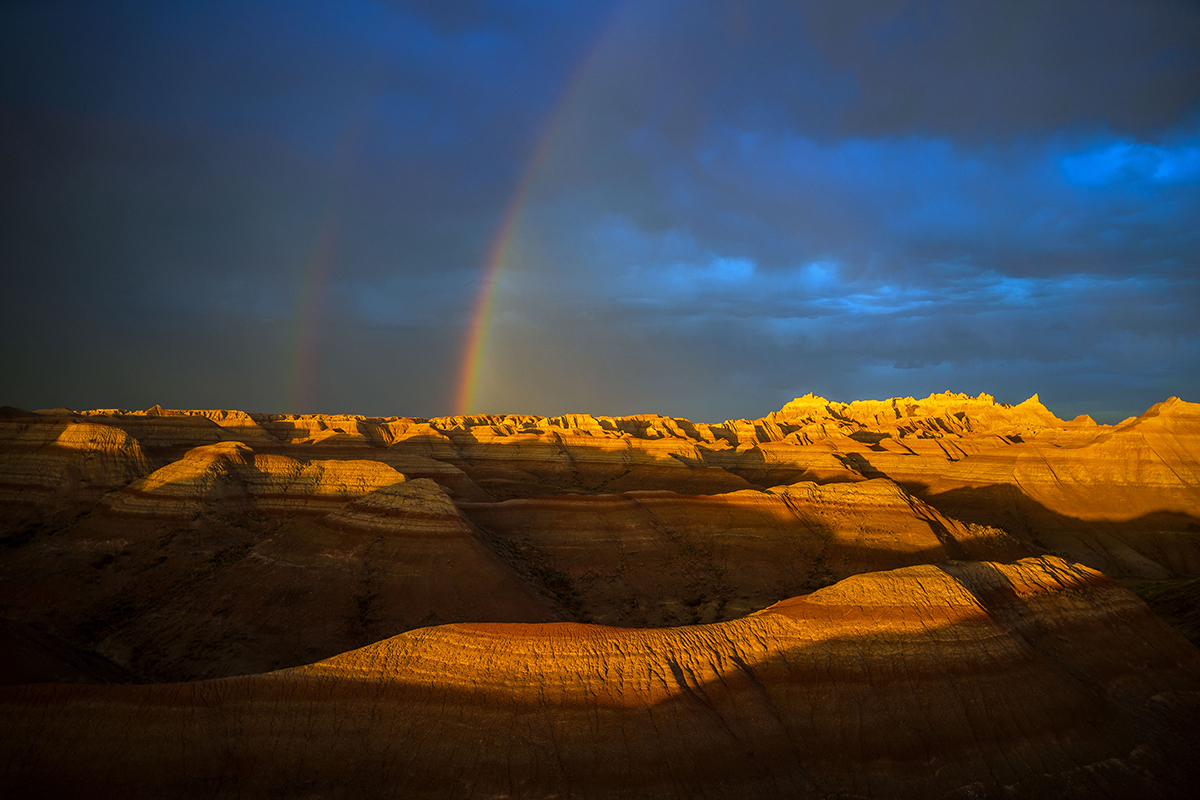 Un espectacular paisaje desértico con formaciones rocosas en capas, iluminado por la luz del sol y con un doble arco iris contra un cielo oscuro, y ejemplo de fotografía de paisaje y meteorología.