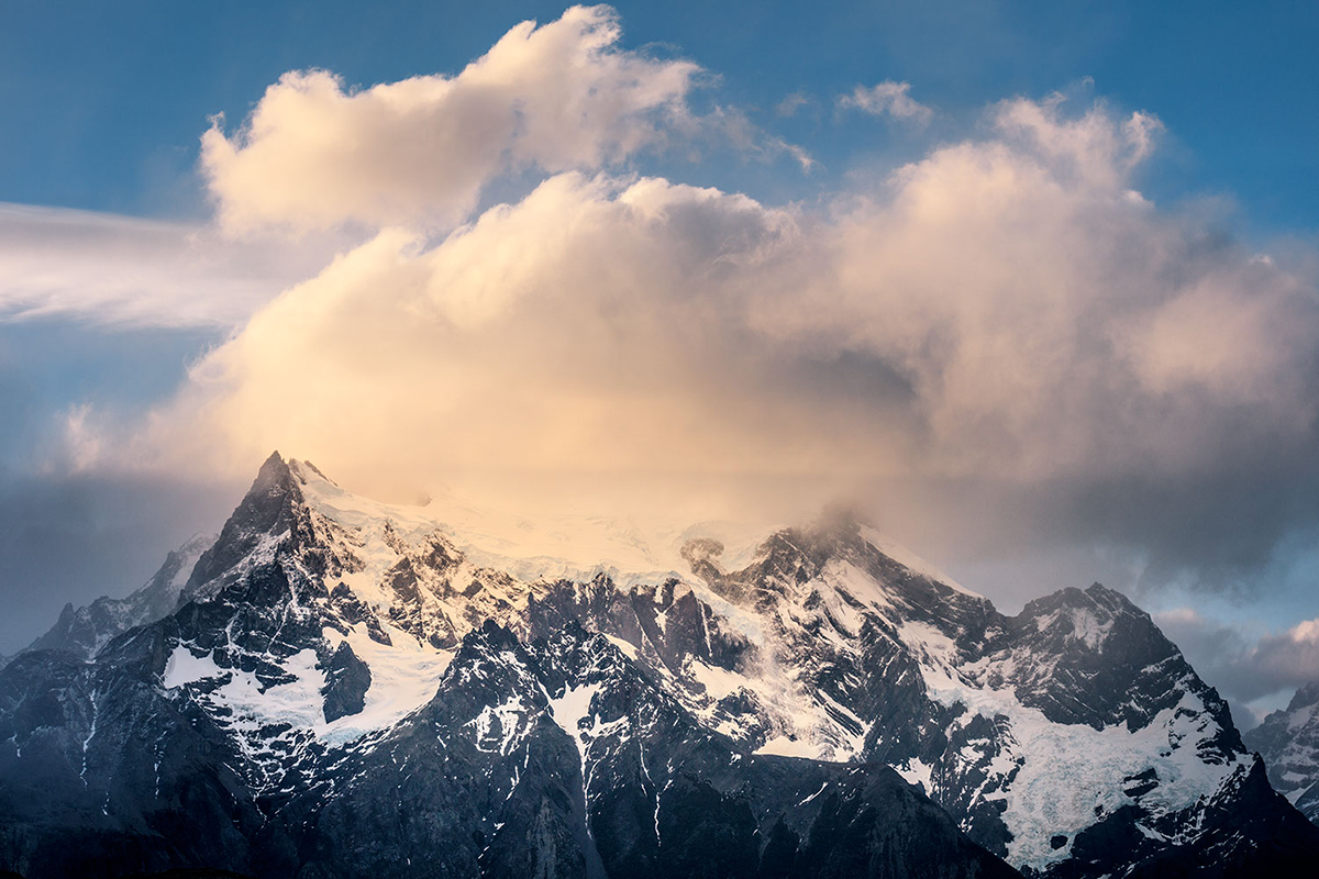 Un pico de montaña cubierto de nieve bajo una nube suave y dorada contra un cielo azul, un gran ejemplo de fotografía paisajística y meteorológica.