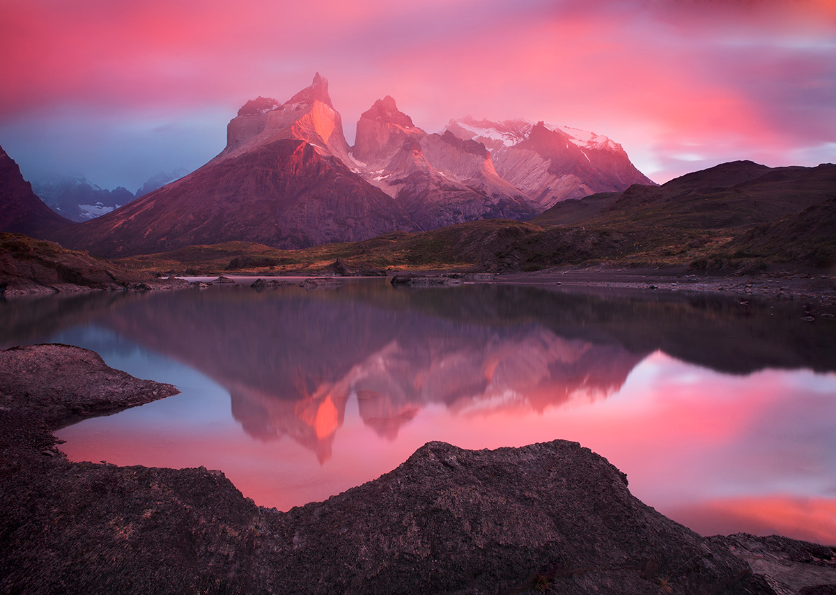 Un sereno paisaje de montaña al amanecer o al atardecer, con cielos rosas y púrpuras reflejándose en un tranquilo lago, rodeado de terreno escarpado, muestra de fotografía de paisaje y meteorológica.