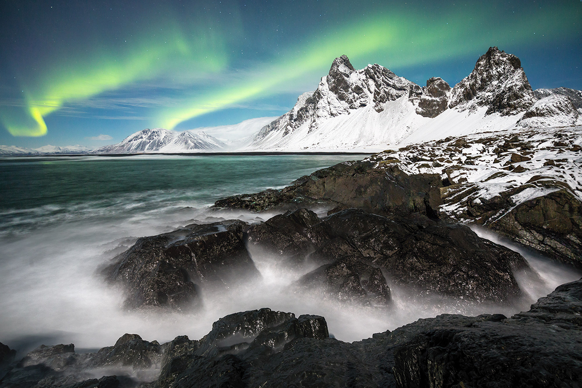 Un paisaje costero nevado con escarpadas rocas negras, mar verde en calma y vibrantes auroras boreales iluminando el cielo. Un bello ejemplo de fotografía paisajística y meteorológica.