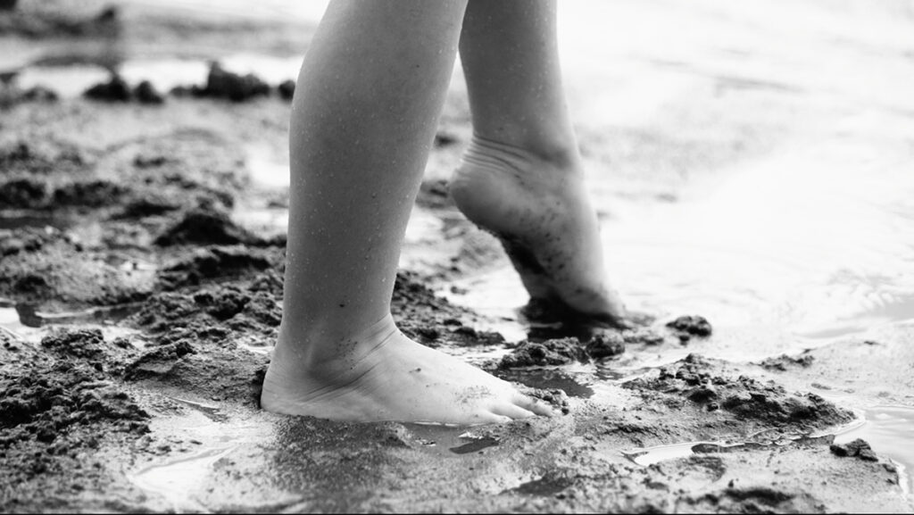 A close-up black and white photograph of a child&rsquo;s bare feet as they walk through wet sand using cinematography lighting techniques to evoke a feeling of carefree exploration.