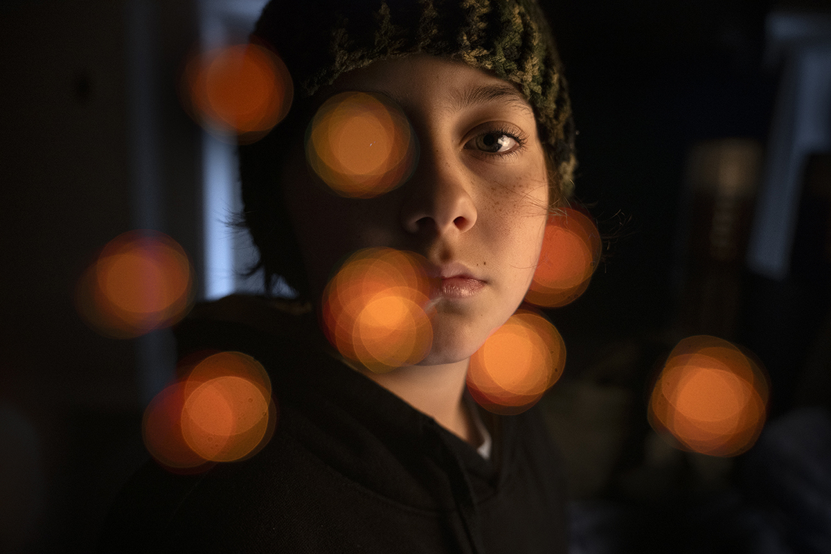Uma criança com um gorro de tricô olha pensativamente, cercada por luzes bokeh laranja quentes e brilhantes.