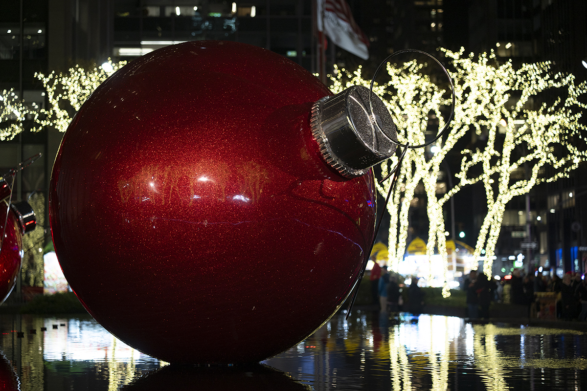 Um enfeite de Natal vermelho gigante reflete as luzes da cidade, cercado por árvores festivas iluminadas à noite.