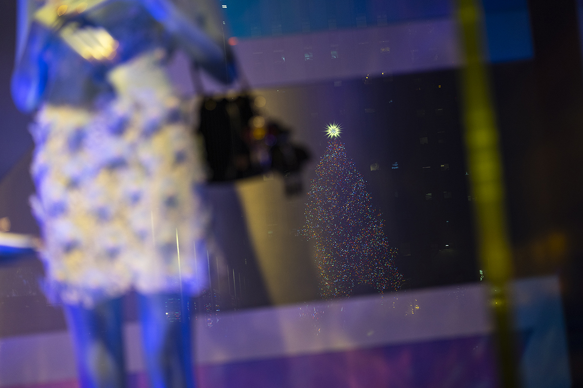 Uma árvore de Natal cintilante se destaca ao longe, refletida na vitrine de uma loja.