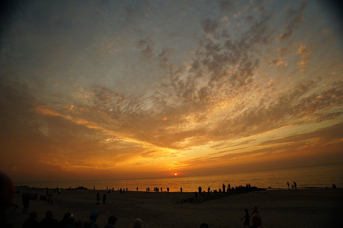 Una vibrante puesta de sol sobre el océano, con siluetas de personas reunidas en la playa, enmarcadas por un cielo lleno de nubes dispersas.