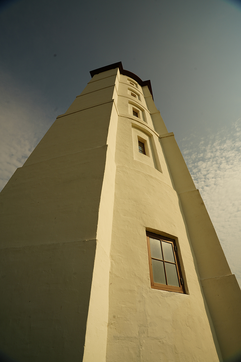 Vista desde un ángulo bajo de un faro alto y blanco con una ventana, sobre un cielo despejado con nubes dispersas.