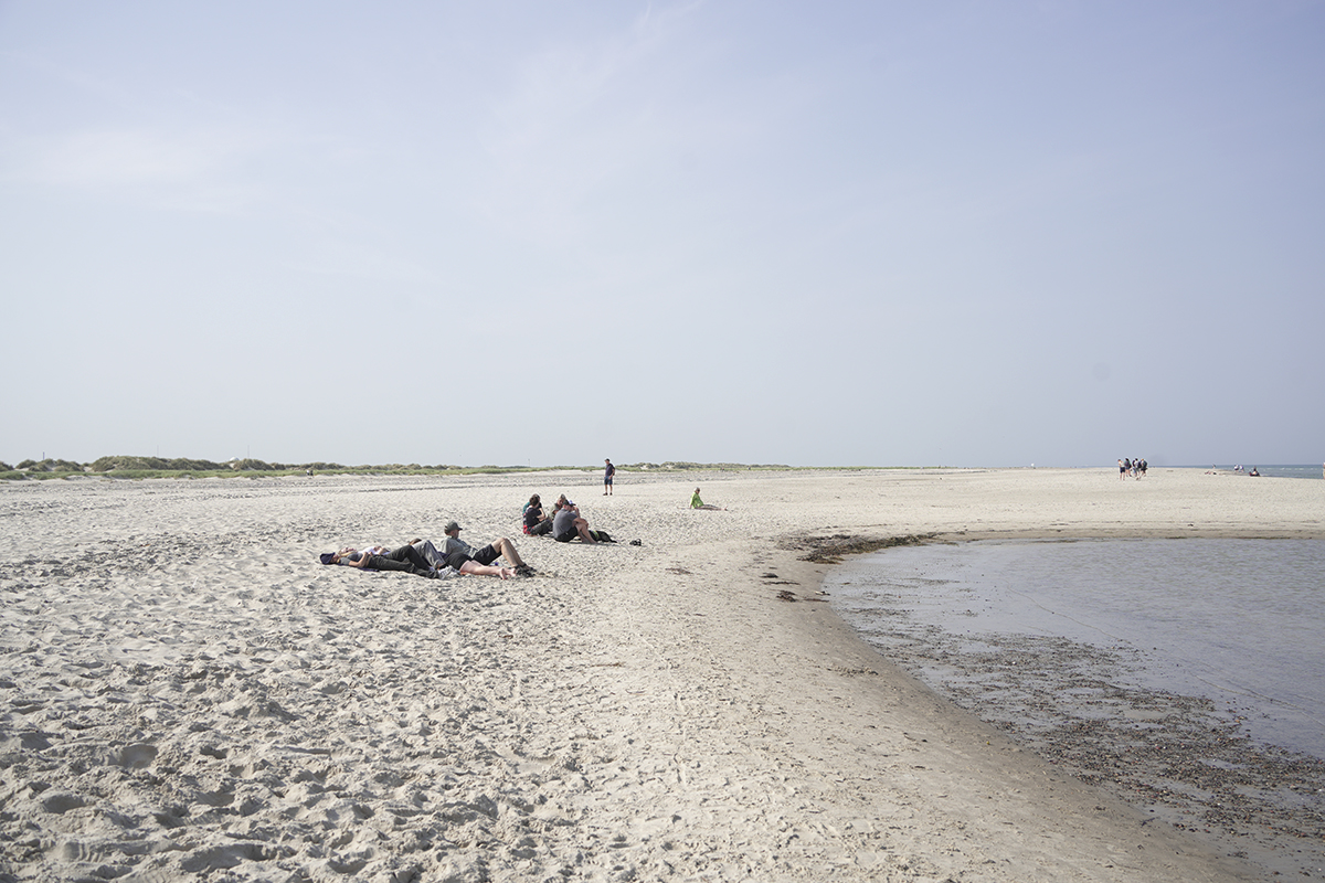 La gente se relaja y se sienta a lo largo de una playa de arena cerca de una orilla poco profunda, con un cielo abierto y dunas lejanas al fondo.