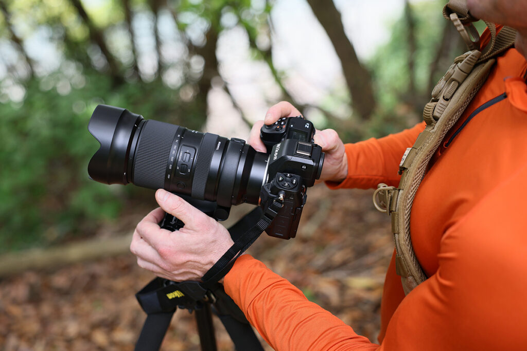 Un photographe en orange tenant un appareil photo monté sur un trépied dans un cadre forestier naturel. 