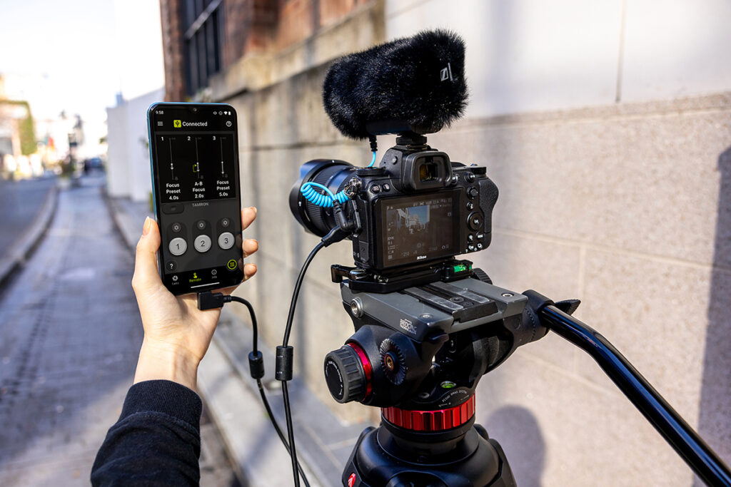A professional camera setup with a camera mounted to a tripod featuring a microphone attachment and connected to a smartphone.