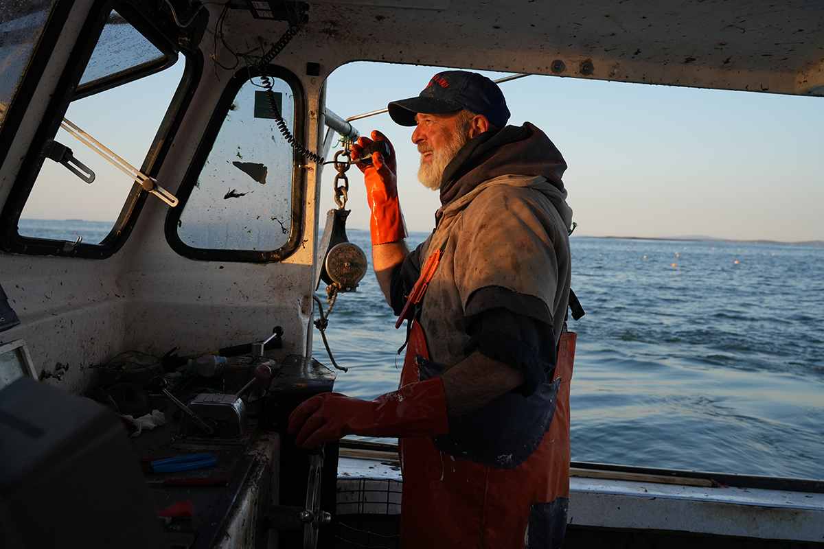 Captured by Rick Gerrity for a feature on photojournalism tips, a seasoned fisherman in orange overalls and gloves operates equipment aboard a fishing boat at sunrise, with the ocean and distant buoys visible through the cabin windows.