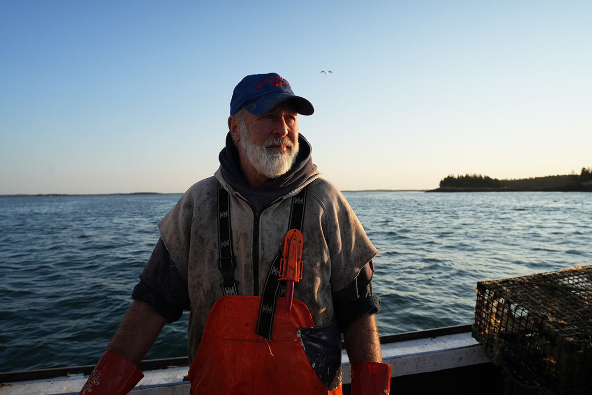 A fisherman in orange overalls stands on his boat at sunrise, gazing thoughtfully over the calm ocean with a lobster trap and a distant shoreline in the background.