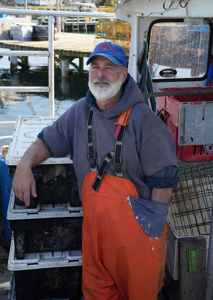 A fisherman in orange overalls and a blue "Beals Island" cap leans on stacked crates aboard his boat, docked near a harbor with lobster traps and fishing equipment in the background. He posed for Rick Gerrity to demonstrate his photojournalism tips.