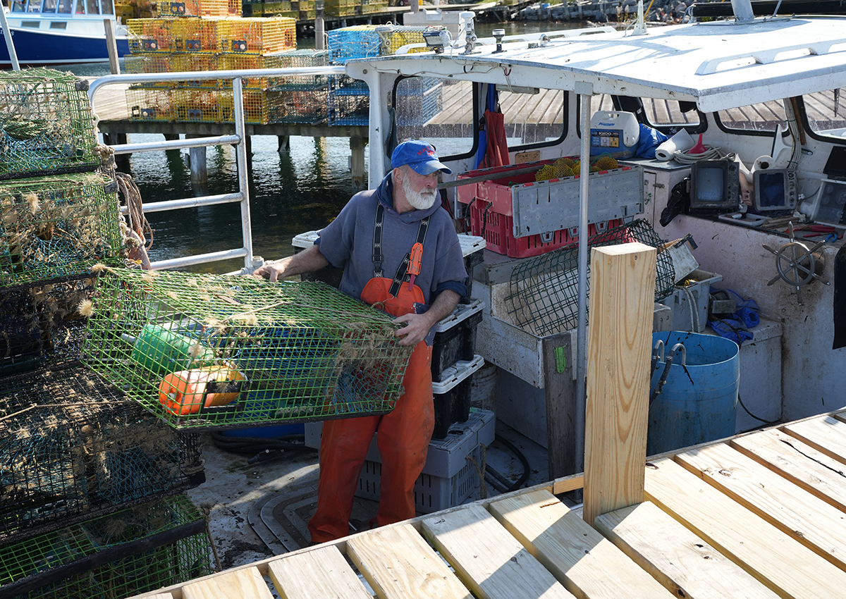 Photojournalism tips in action: A fisherman in orange overalls unloads a green lobster trap from his boat onto a dock, surrounded by stacked traps and harbor equipment in a bustling waterfront setting.