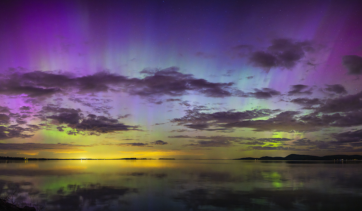 A vibrant display of the Northern Lights in purple, green, and yellow hues reflecting over a calm lake at twilight&mdash;demonstrating how to photograph the northern lights in transitional light conditions with balanced exposure.