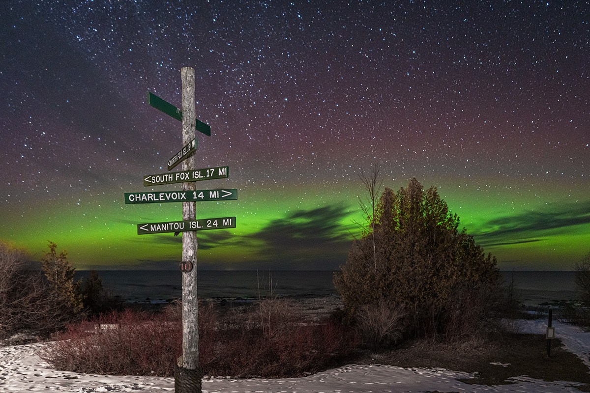 A rustic wooden signpost under a vivid aurora-lit sky, with snow-covered ground and trees in the foreground.