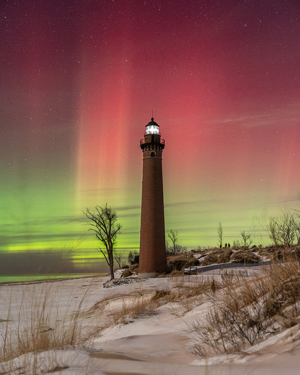 A tall lighthouse illuminated against a vibrant red and green aurora, with a snowy beach and bare trees in the foreground.