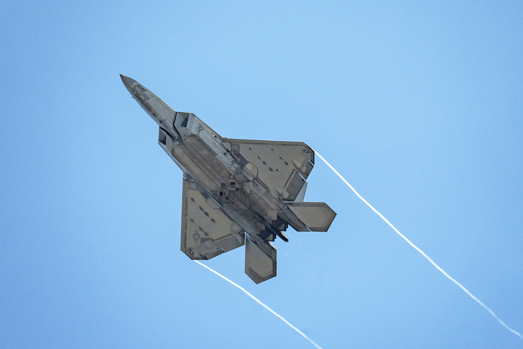 Un F-22 Raptor capturado desde abajo, dejando finas estelas de vapor contra un cielo azul despejado.