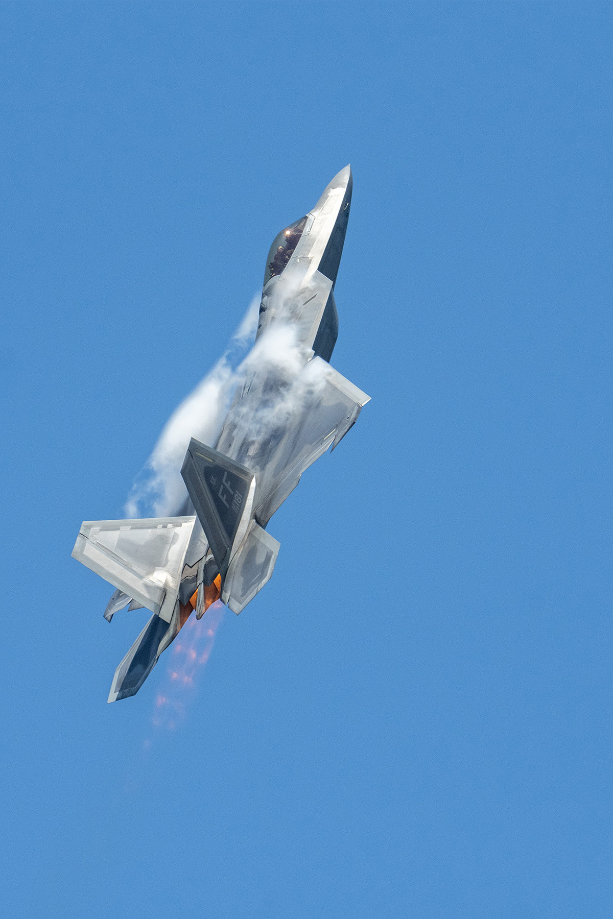 Un F-22 Raptor asciende bruscamente con los postquemadores encendidos y las estelas de vapor formándose contra un cielo azul brillante, demostrando técnicas expertas de fotografía de espectáculos aéreos.