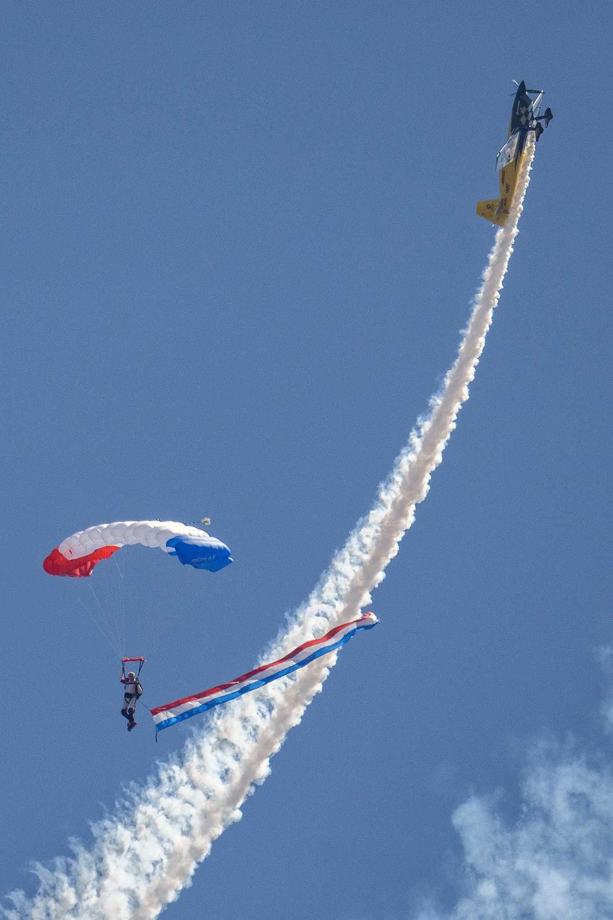Un paracaidista con una colorida campana desciende mientras un avión acrobático asciende, dejando una estela de humo en el cielo azul despejado. Lee las técnicas de fotografía de espectáculos aéreos de Tamron para aprender a captar la estela de vapor.