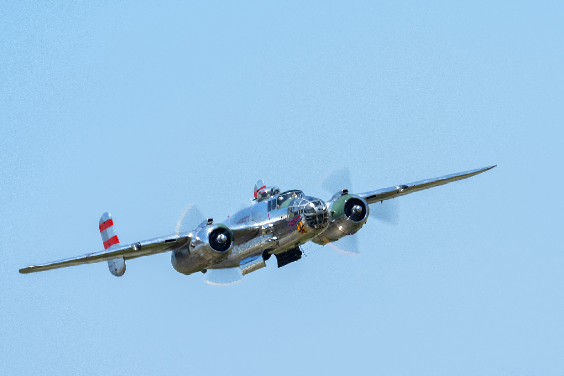 Un avión plateado de época con hélices giratorias capturado en pleno vuelo contra un cielo azul despejado.