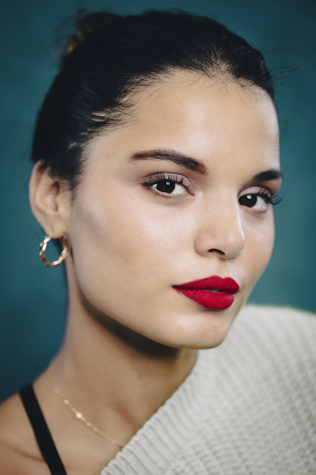 Retrato en primer plano de una mujer con un llamativo pintalabios rojo, pendientes de aro dorados y una mirada segura sobre un fondo verde azulado, capturado con uno de los mejores objetivos Nikon Z para fotografía de retrato.