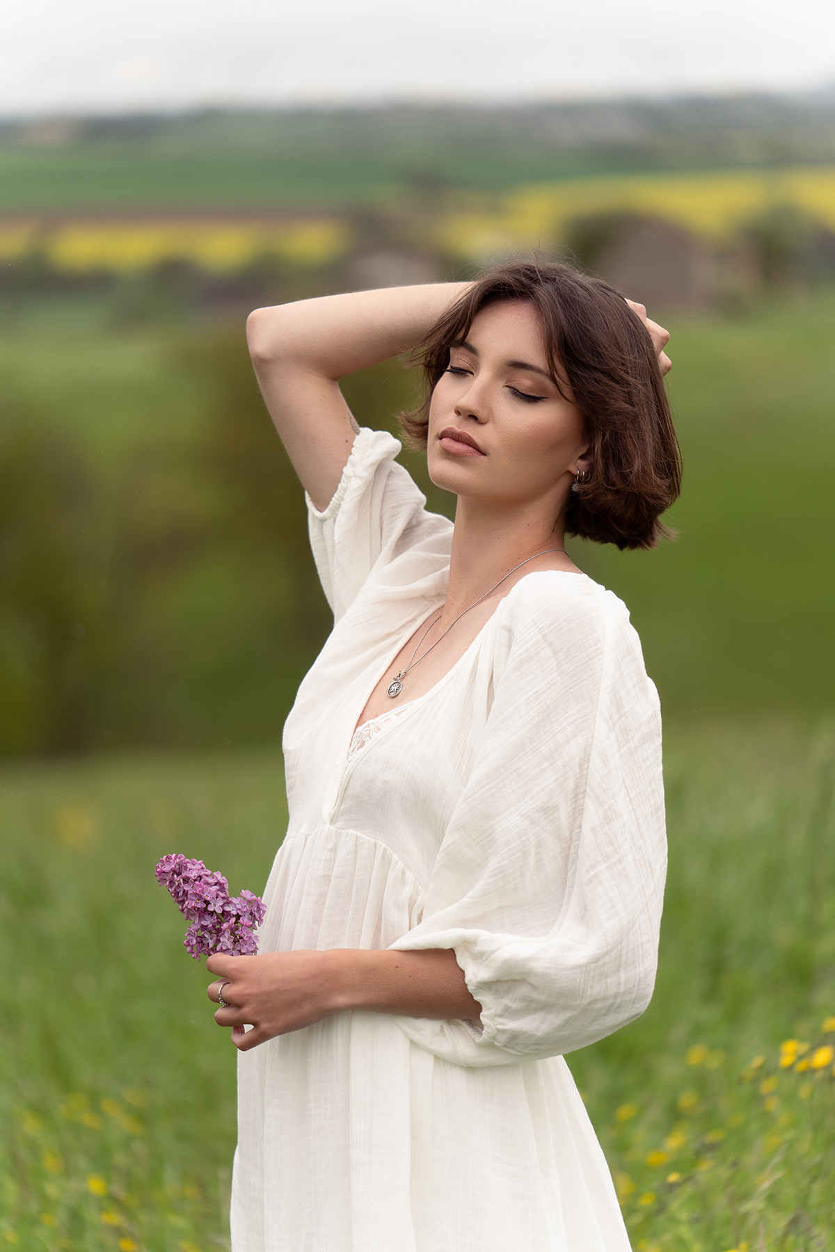 Une jeune femme vêtue d'une robe blanche fluide et tenant des fleurs violettes, debout dans un champ verdoyant, avec une expression sereine. Cette photo a été prise à l'aide des meilleurs objectifs Sony à monture E pour la photographie de portrait.