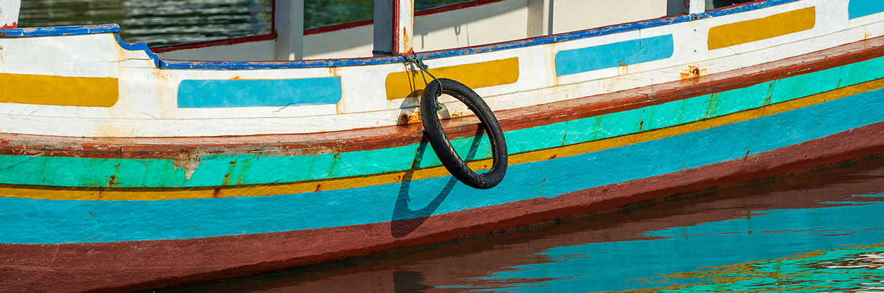A colorful wooden boat with a hanging tire reflecting on calm water.