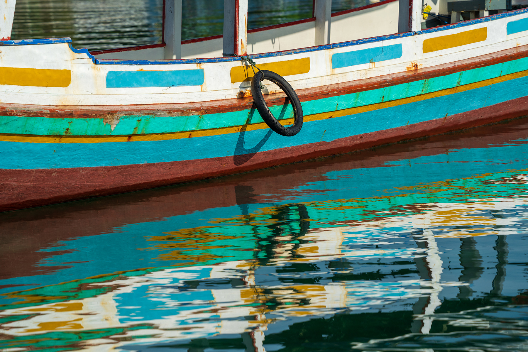 A vibrant wooden boat with a hanging tire reflecting on the rippling water.