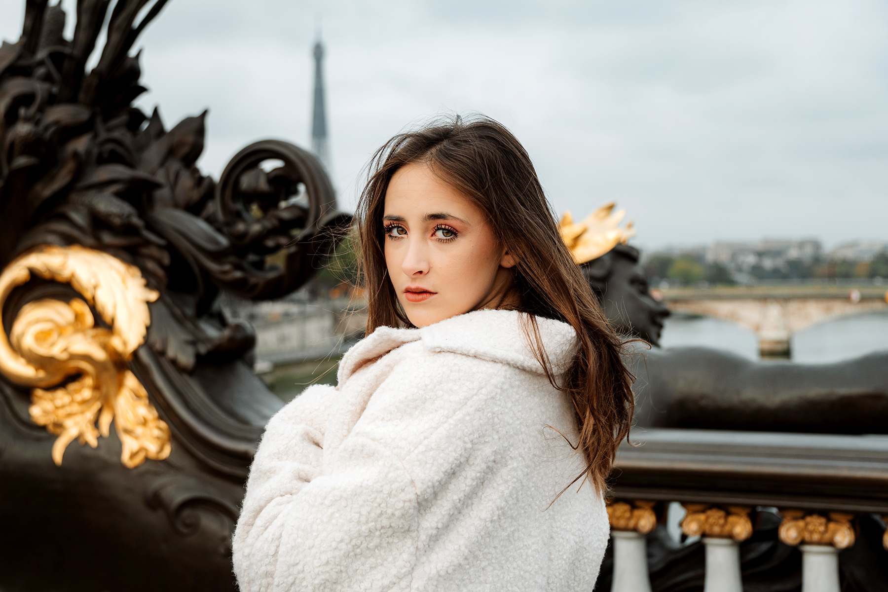 A woman in a white coat poses elegantly on a bridge with ornate details, the Eiffel Tower visible in the background, photographed with the Tamron 17-70mm E-mount APC-C lenses.