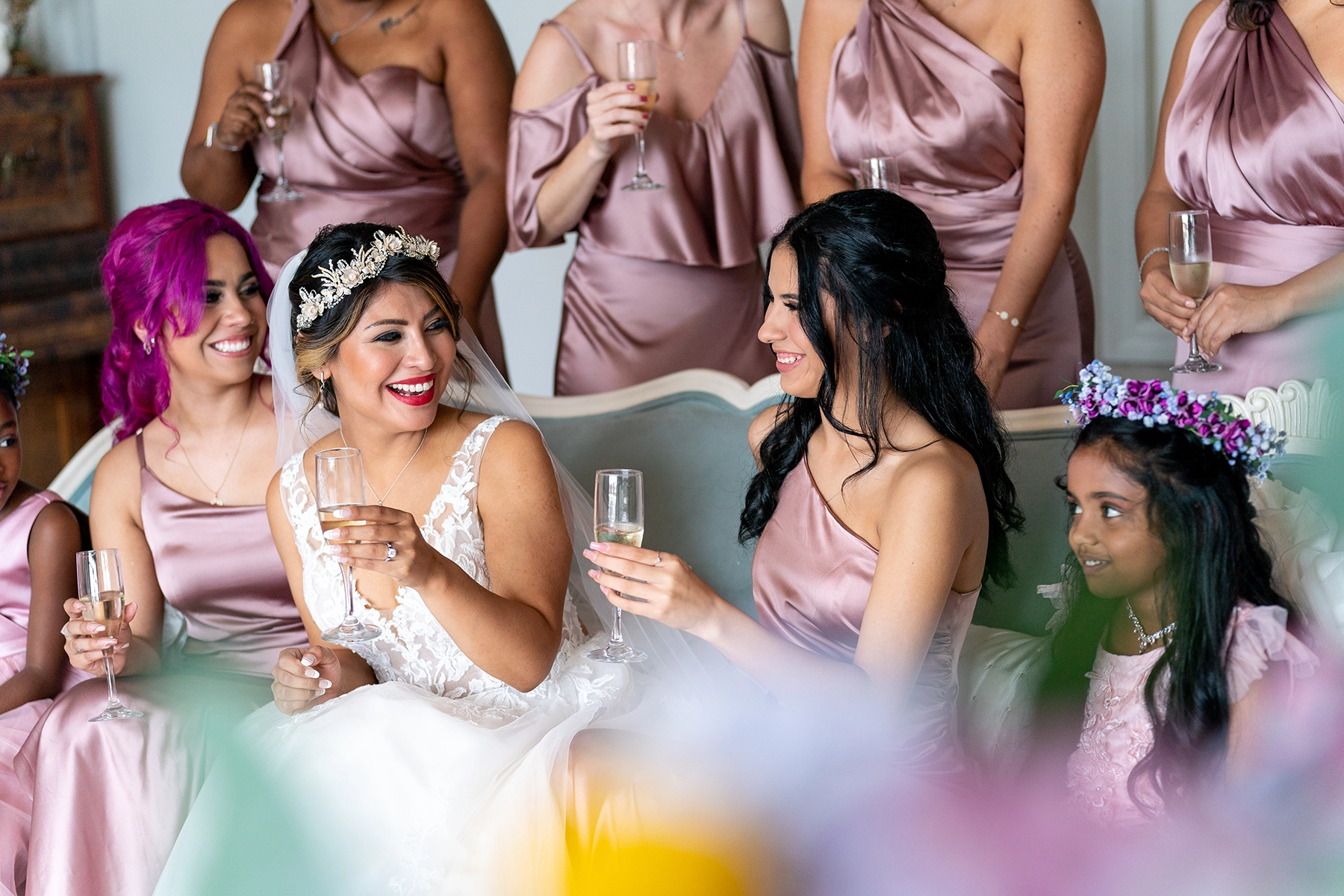 La novia ríe con las damas de honor y la niña de las flores, vestidas con trajes de satén rubor, mientras brindan con champán antes de la ceremonia nupcial. Una imagen de fotografía de boda de estilo documental, que captura un momento de alegría y celebración con un enfoque refinado y narrativo.