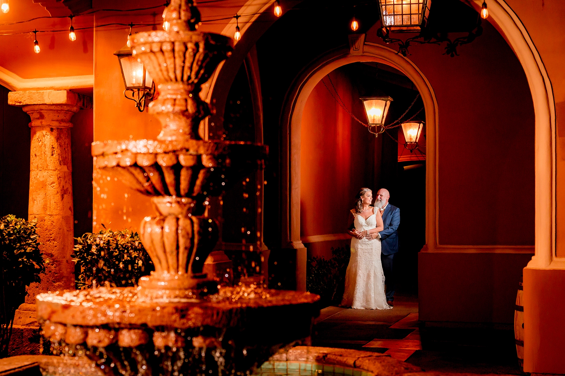 Un elegante retrato editorial de fotografía de boda de una pareja de recién casados posando bajo una cálida iluminación ambiental cerca de una gran fuente en un exclusivo lugar de celebración de bodas. La novia, con un vestido de encaje, y el novio, con un traje a medida, comparten un íntimo abrazo, enmarcados por arcos arquitectónicos y faroles brillantes, creando una estética cinematográfica de alta costura.