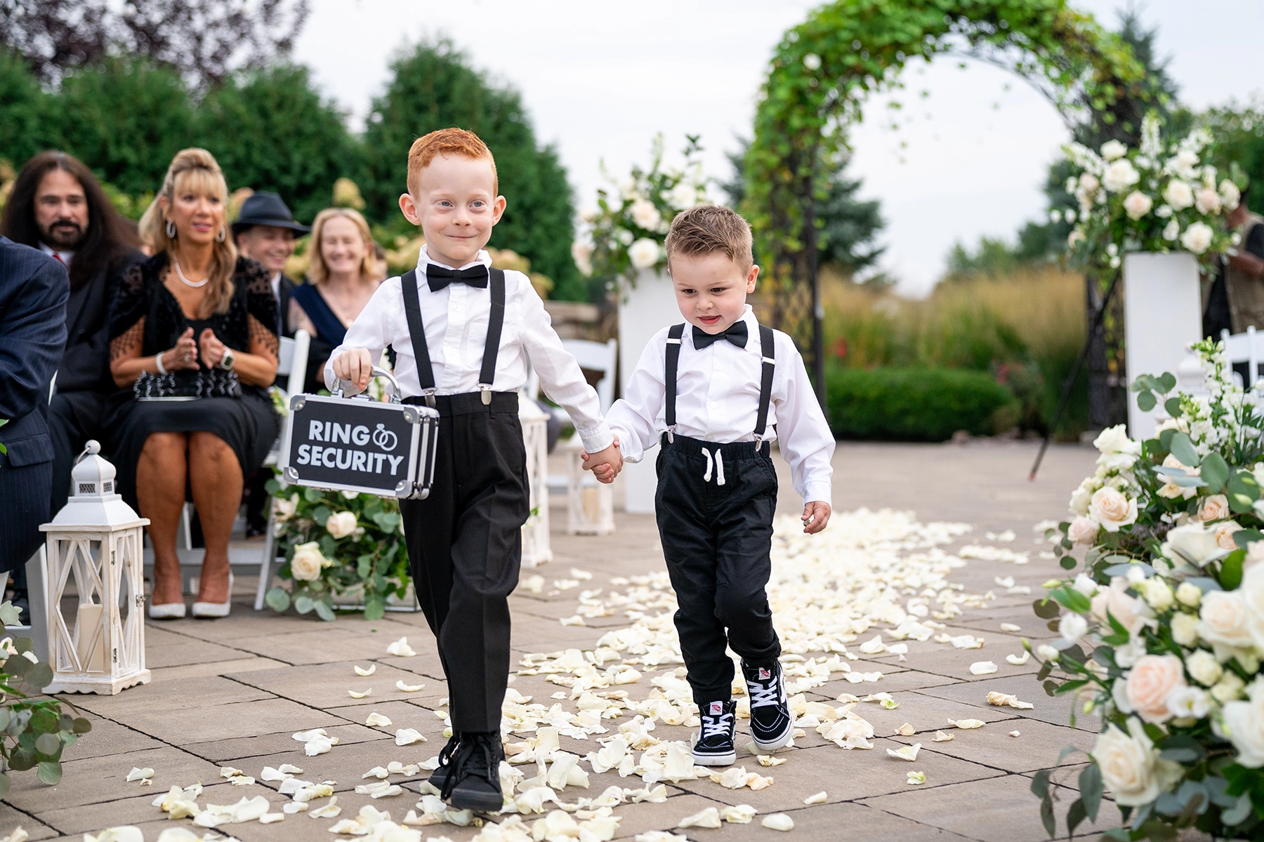 Two young ring bearers walk down the aisle holding hands, one carrying a "Ring Security" briefcase, beautifully captured in documentary style wedding photography.