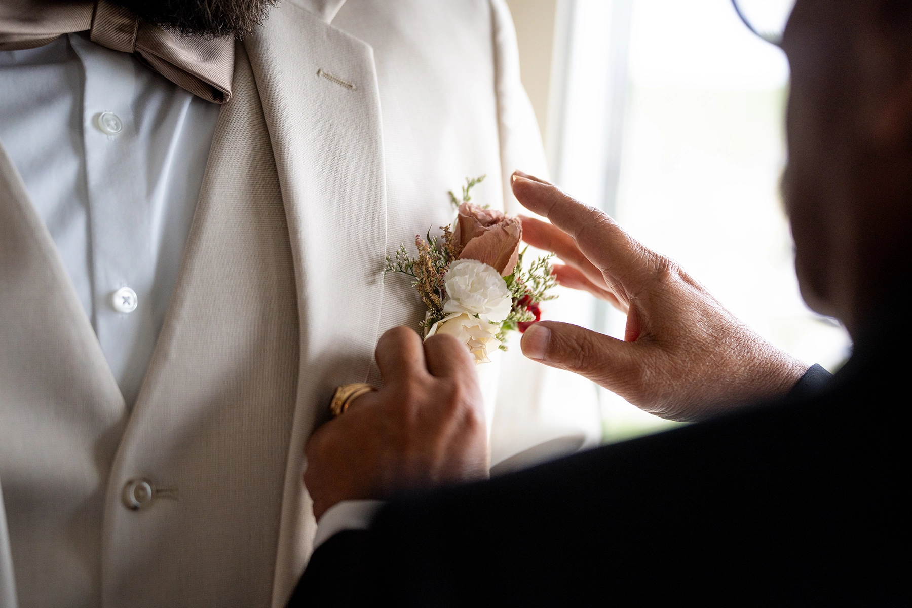 A close-up candid moment of a groom having his boutonniere adjusted, captured in documentary style wedding photography with natural light and soft focus.
