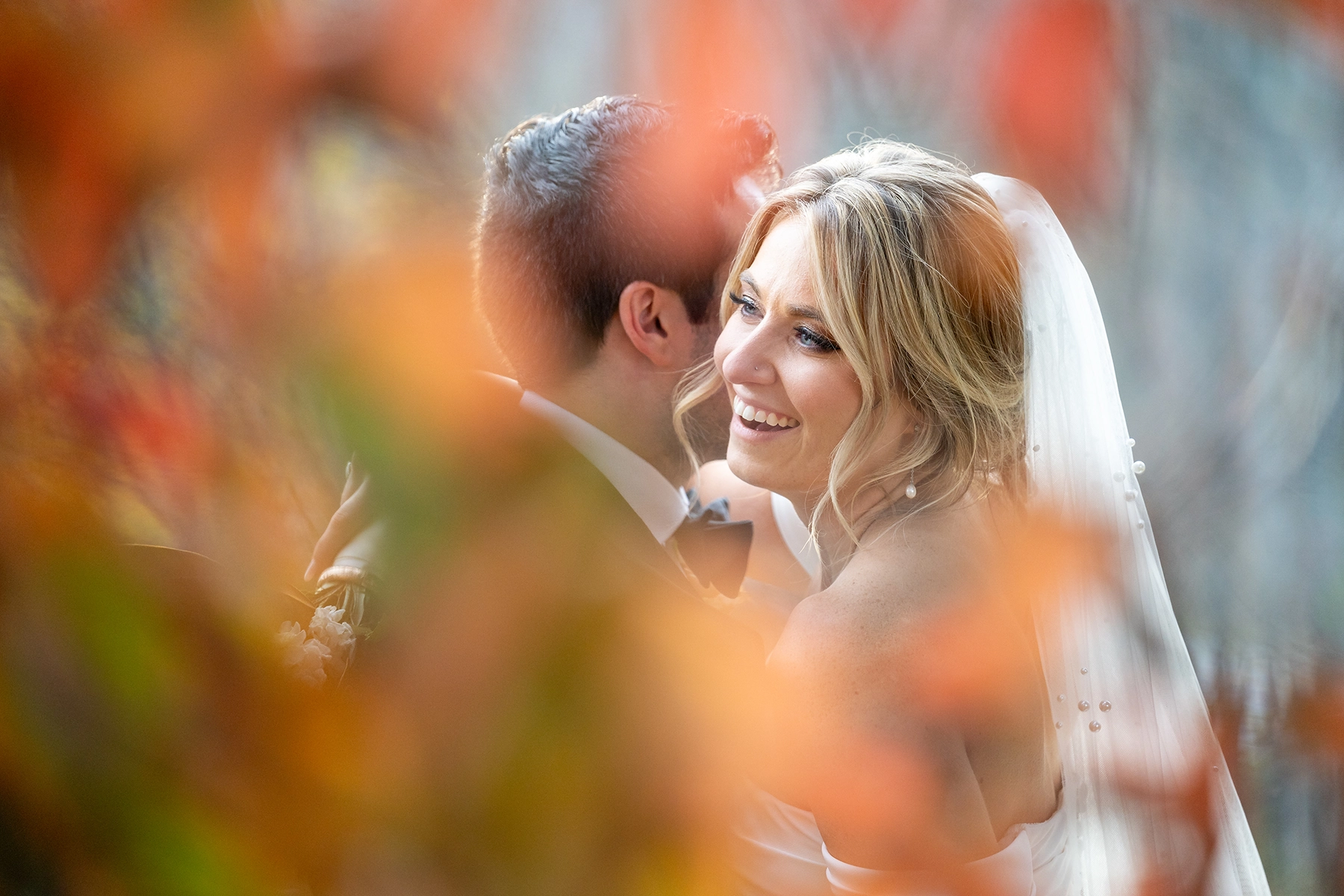 Una novia y su novio comparten un momento íntimo, capturado a través del follaje otoñal. La novia, radiante de felicidad, abraza a su pareja, reflejando las tendencias en fotografía de bodas de 2025 con una mezcla de narración documental y suave elegancia editorial.