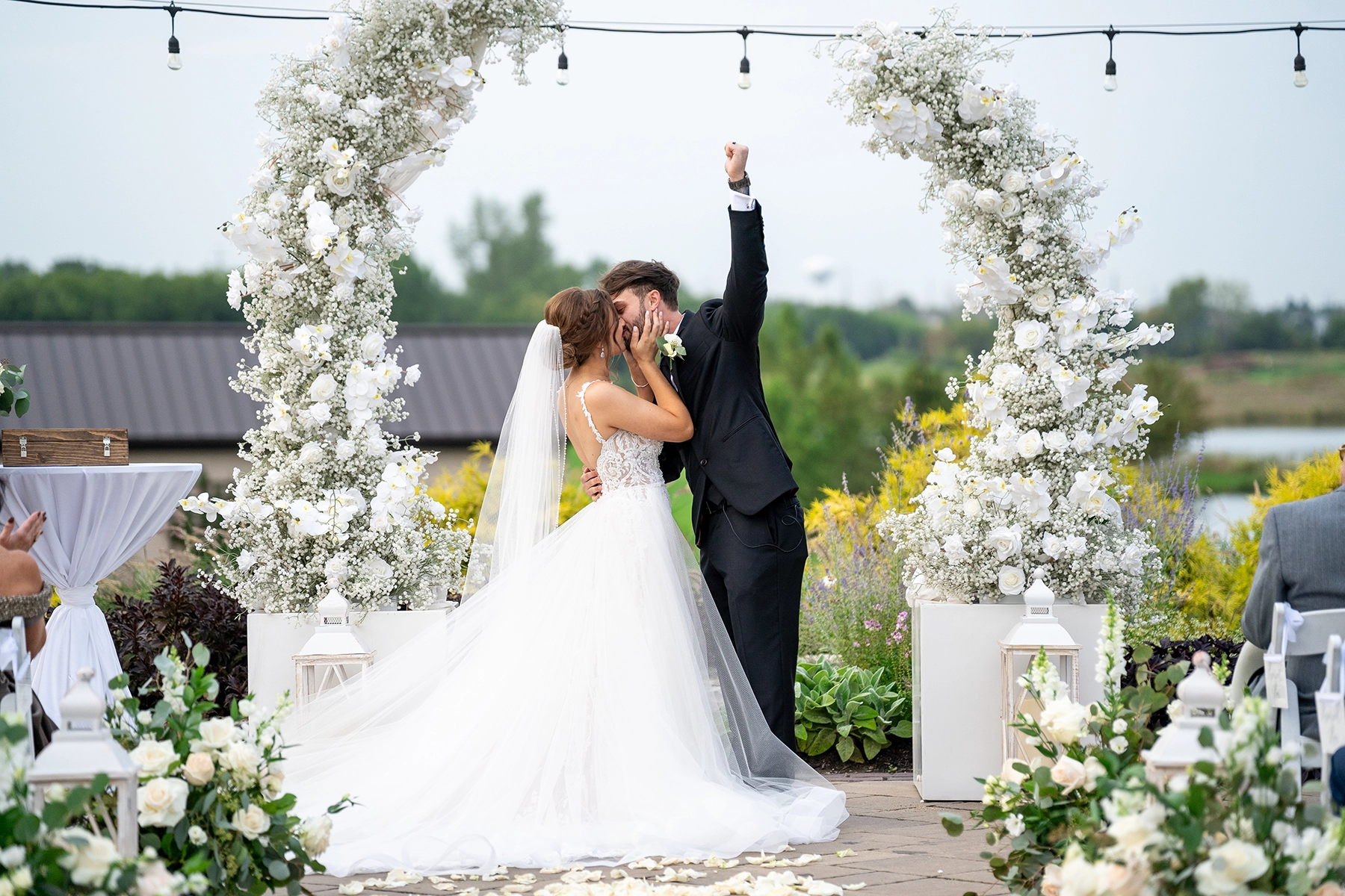 Una pareja de recién casados comparte su primer beso bajo un impresionante arco floral, con el novio levantando el puño en señal de celebración. La ceremonia al aire libre, enmarcada por exuberantes flores blancas y farolillos luminosos, encarna la esencia de las tendencias en fotografía de bodas de 2025 con un estilo cinematográfico y narrativo.