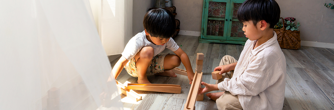 Dos niños pequeños juegan con bloques de madera en el suelo en una habitación iluminada de forma natural, con una suave luz solar que entra por una ventana cubierta por cortinas transparentes.