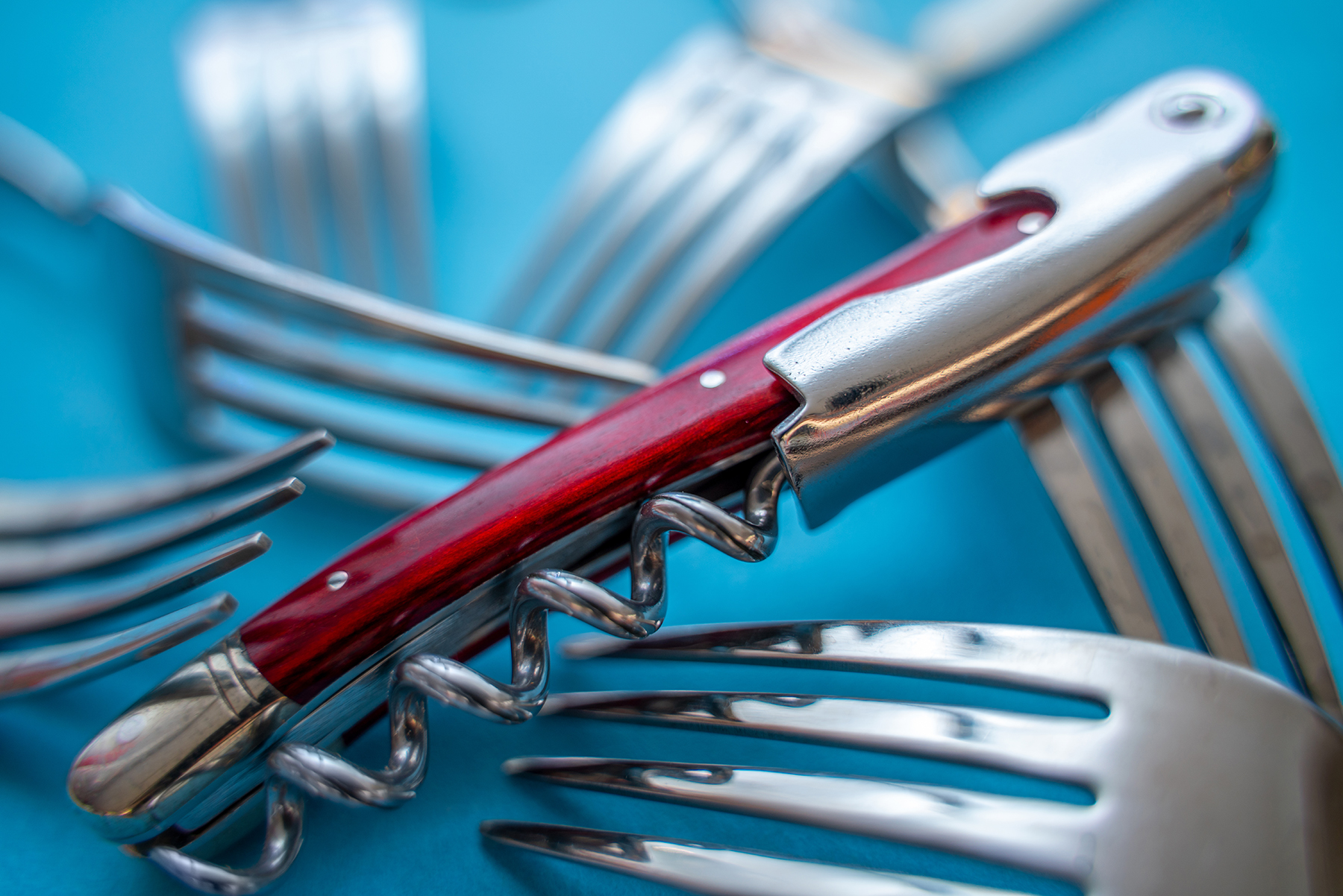 Primer plano de un sacacorchos con mango rojo rodeado de tenedores de acero inoxidable superpuestos sobre un fondo azul brillante, creando una composición dinámica y reflectante.