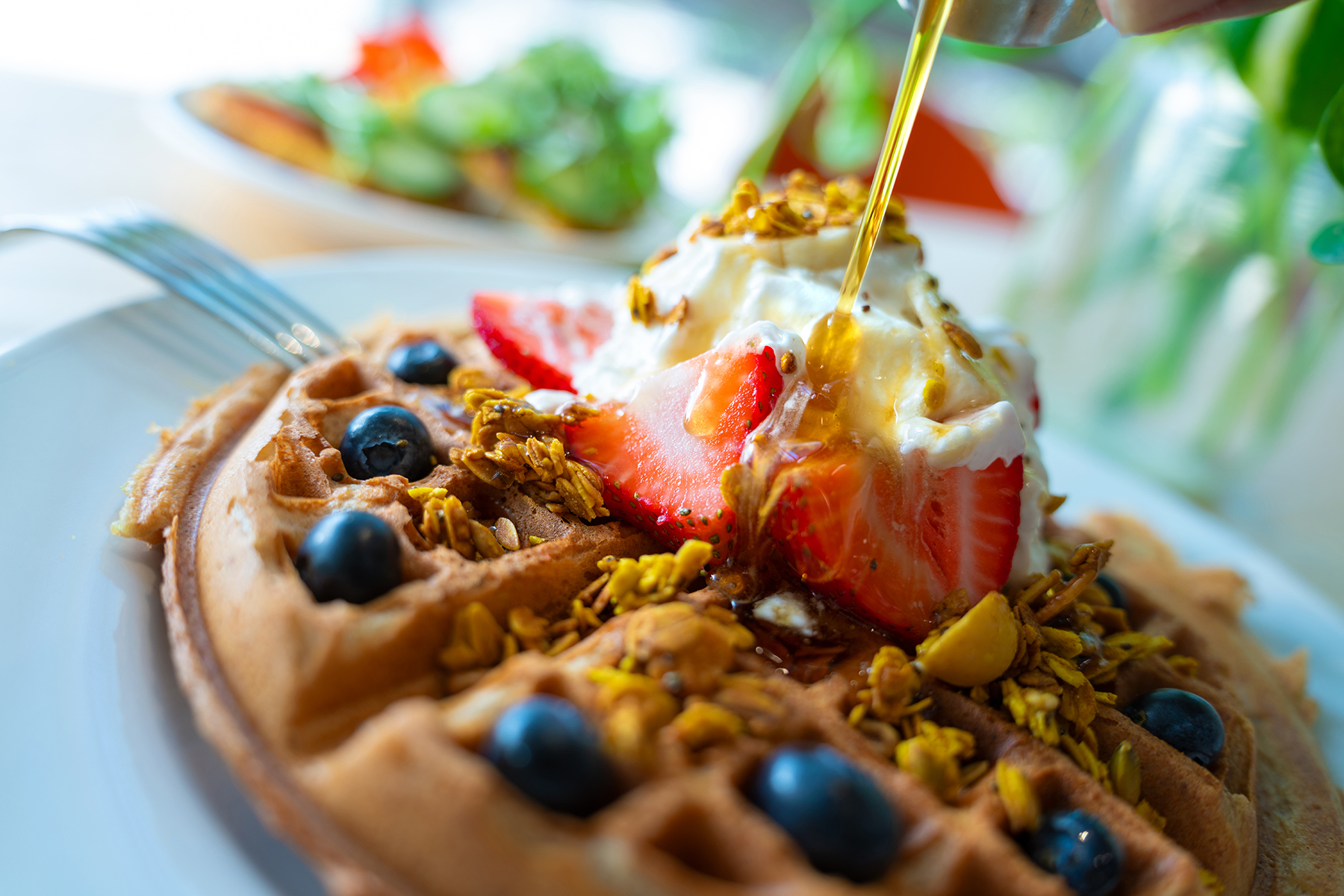 Un primer plano de un gofre cubierto con fresas frescas, arándanos, granola, nata montada y un chorrito de sirope dorado, con un fondo difuminado en el que se ve vegetación vibrante y otros platos de comida.