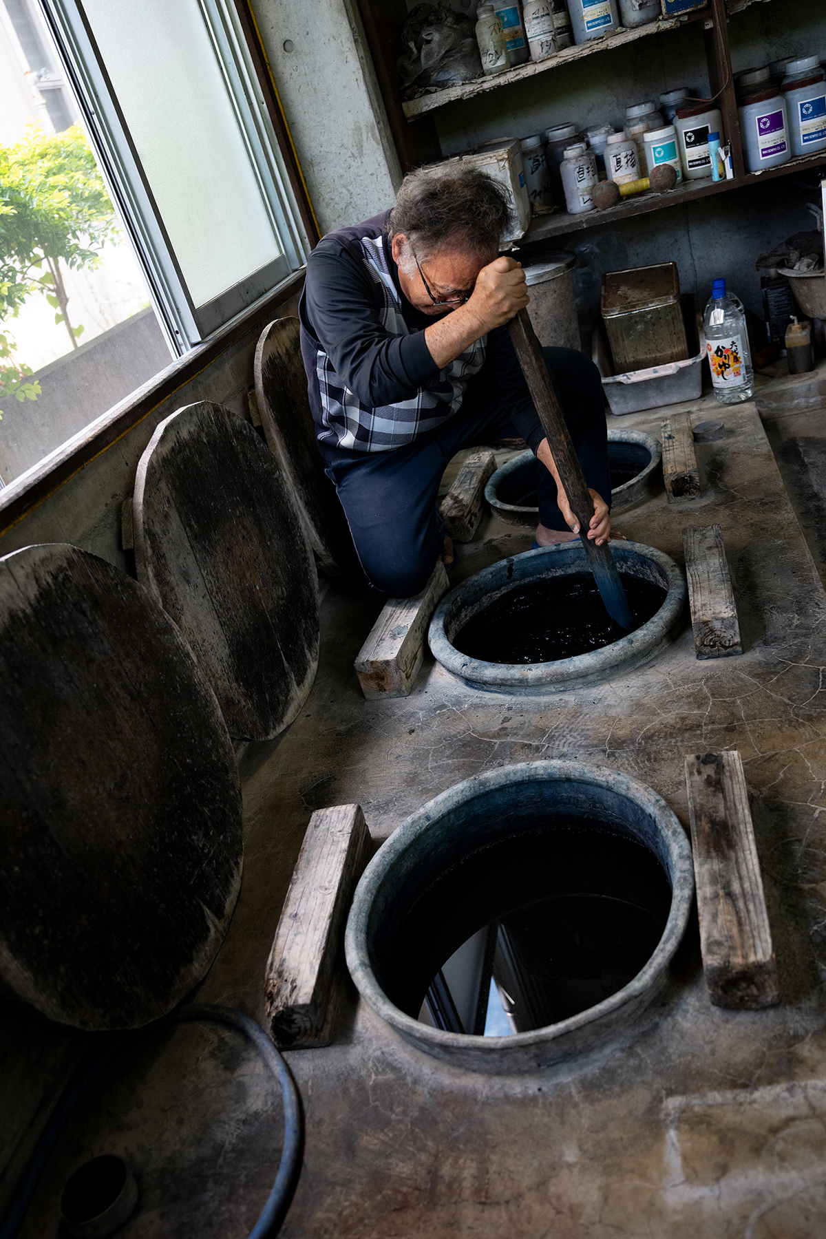 Un artesano remueve con cuidado grandes cubas de añil en un taller rústico. La luz natural entra por una ventana cercana e ilumina el espacio de trabajo de madera y hormigón lleno de herramientas y materiales.