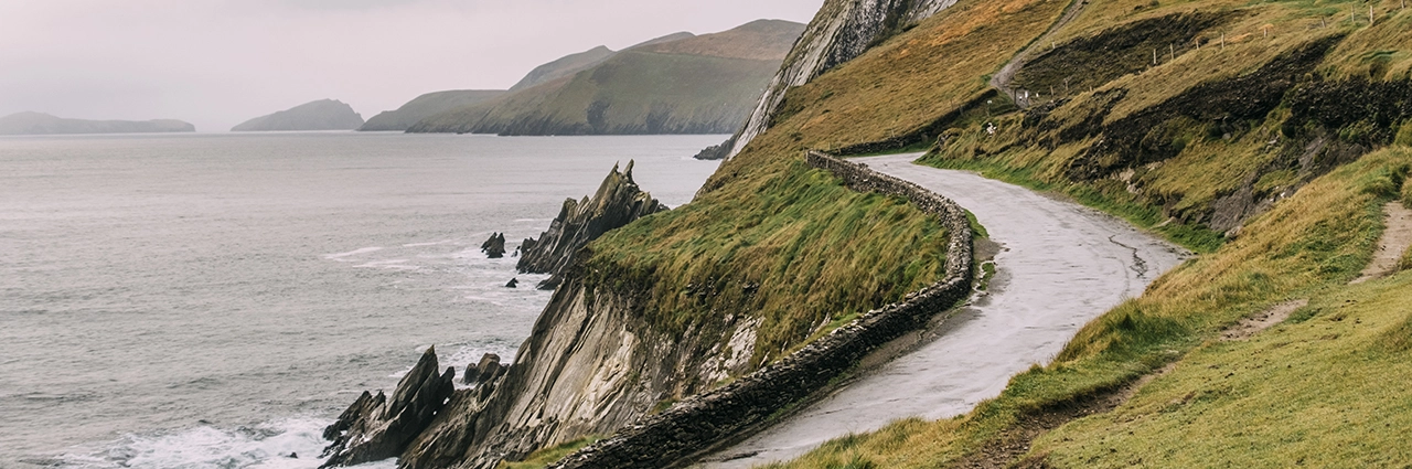 Scenic coastal road winding along rugged cliffs and the ocean under overcast skies, captured with a wide-angle lens—ideal for showcasing depth and dramatic landscapes.