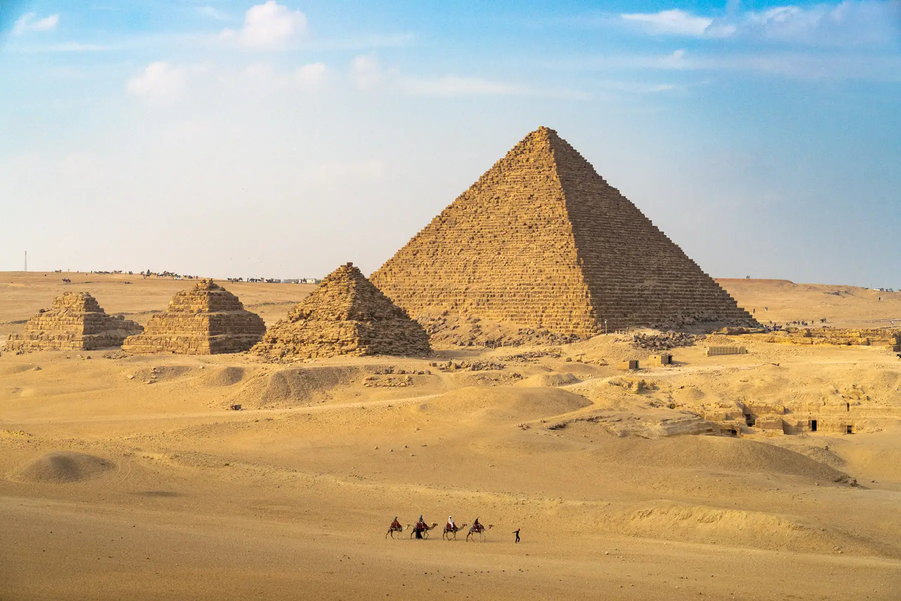 Camel riders travel across the desert in front of the Pyramid of Menkaure and smaller tombs at Giza, a stunning scene to photograph historical landmarks in Egypt.
